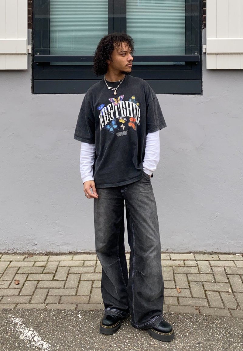 Fekete, túlságosan nagy grafikai póló, színes pillangó mintával, fehér hosszúujjú póló felett, laza sötét farmernadrággal és vastag fekete cipőkkel párosítva.