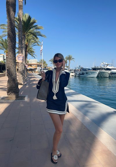 Vestido de marina con acentos blancos y un cuello, que presenta un fleco en la parte delantera. Lleva un bolso tejido en blanco y negro, y sandalias negras. Palmeras y barcos en el fondo.