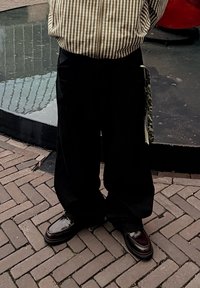 Černé široké kalhoty v kombinaci s lesklými hnědými koženými mokasíny. Celý outfit doplňuje pruhovaná hnědo-bílá košile s knoflíky.