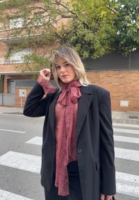 Γυναίκα που στέκεται σε διάβαση πεζών φορώντας μαύρο σακάκι και διάφανο βυσσινί δαντελένιο μπλουζάκι με μεγάλο φιόγκο, διορθώνοντας τα μαλλιά της, με κτίρια από τούβλα και δέντρα στο βάθος.