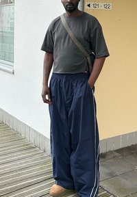 Pantalon ample bleu marine avec des bandes blanches sur les côtés, associé à une courte chemise gris foncé. La tenue comprend des chaussures couleur fauve et un sac porté en bandoulière.