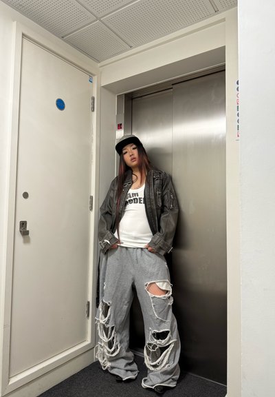 Pantalones de chándal grises de gran tamaño con grandes rotos, camiseta sin mangas blanca con el texto "I AM MODELED" y una chaqueta de cuero negra; gorra negra. Fondo de ascensor.