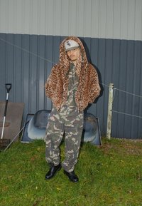 Jakna iz umetnega krzna z vzorcem leoparda in kapuco, olivno zelena kamuflažna kombineza, črni čevlji, postavljeni ob sivi steni in vrtnih orodjih na travi.