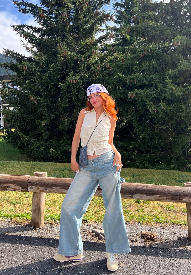 Λευκή αμάνικη μπλούζα με πτυχωτή υφή, ανοιχτό μπλε φαρδιά τζιν παντελόνια, λευκά sneakers με χρωματικές λεπτομέρειες και ένα μοτίβο μωβ μαντήλι.