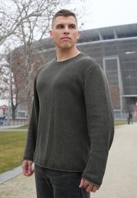 Pull à col rond en vert foncé, motif en maille texturée, manches longues et coupe décontractée. Associé à un pantalon gris foncé.