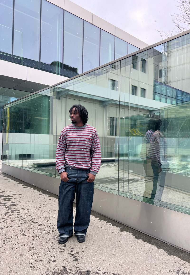 Homme aux dreadlocks portant un pull rayé et un jean large, debout près d'un bâtiment en verre, reflétant son image sur la surface vitrée.