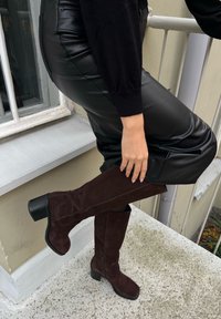 Botas de camurça castanhas até ao joelho com salto quadrado e fecho, combinadas com calças de couro pretas, exibidas sobre uma superfície de pedra.