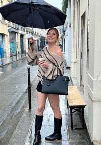 Strickpullover mit cremefarbenen und schwarzen Streifen, schwarze Handtasche, schwarze kniehohe Stiefel, hält eine Kaffeetasse, unter einem schwarzen Regenschirm, auf einer nassen Straße.