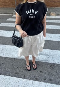 Melns un balts svītrots Nike T-krekls ar īsām piedurknēm, baltas šortus ar auklu, melna plecu soma un melni flip-flopi.