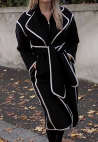 Black trench coat with white piping, featuring a tie waist, side pockets, and a double-breasted design. Textured fabric, knee-length.