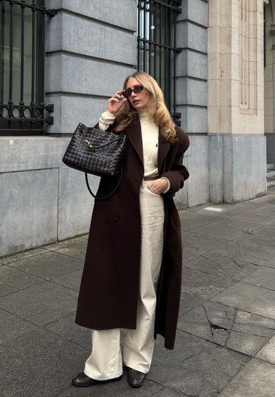 Abrigo de lana marrón, suéter de punto color crema, pantalones beige claros, zapatos de cuero marrón. Bolso de mano tejido negro, gafas de sol oscuras. Fondo urbano.