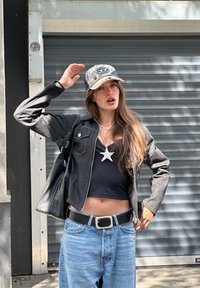 Čierna skrátená bunda, čierny top bez rukávov, svetlo modré voľné džínsy, strieborný prívesok hviezdy. Čiapka trucker s logom, čierna kabelka cez rameno, šedé pozadie.