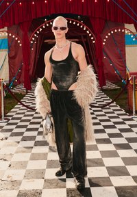 Μαύρο βελούδινο κορμάκι, ταιριαστά παντελόνια και παλτό από τεχνητό γούνα. Τα αξεσουάρ περιλαμβάνουν ασημένια κοσμήματα και γυαλιά ηλίου. Το φόντο απεικονίζει την είσοδο του τσίρκου.