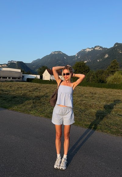 Top corto de rayas en azul claro y pantalones cortos de algodón. Zapatillas blancas con suela más gruesa. Bolsa de hombro marrón con estampado. Fondo de paisaje natural.