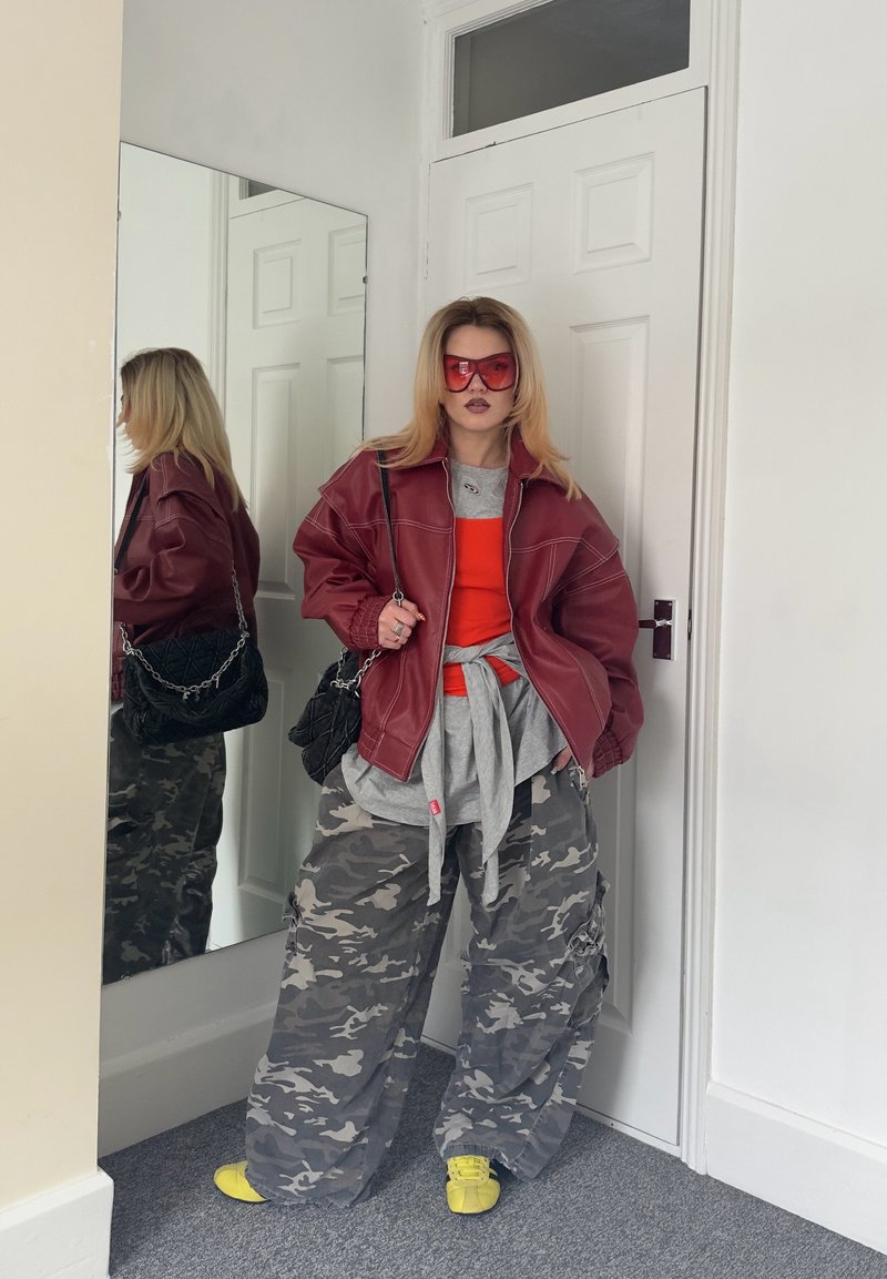 Personne portant une veste bordeaux oversize, un haut orange, un pantalon camouflage, des chaussures jaunes et des lunettes de soleil rouges, debout près d'une porte avec un miroir reflétant leur profil.