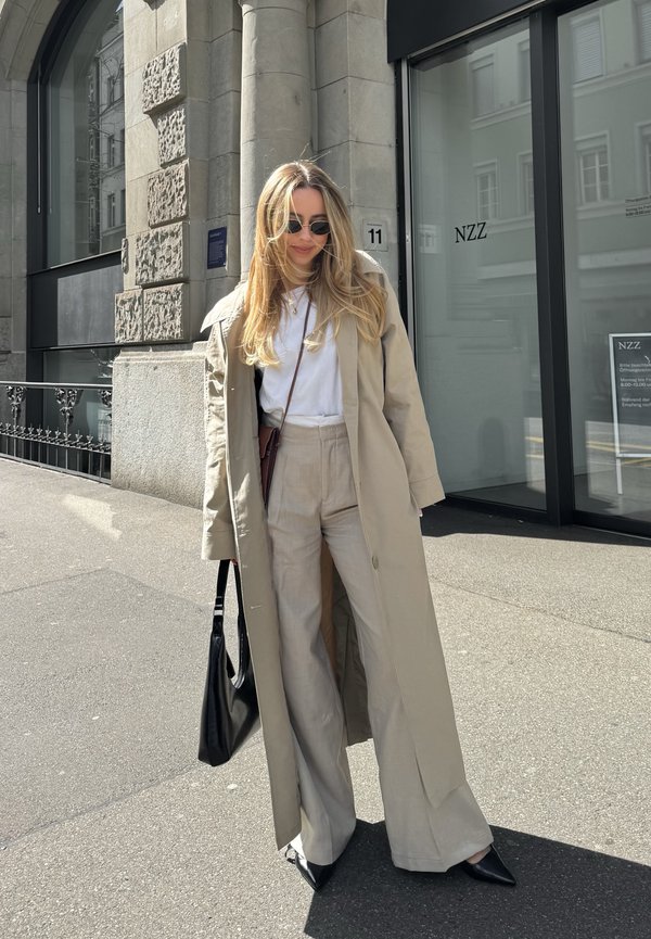 Un trench coat bej peste o cămașă albă, asortat cu pantaloni largi bej deschis. O geantă de mână neagră și tocuri negre cu vârf ascuțit completează ținuta.