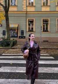 Brown leather trench coat with a belted waist, teamed with a light pink jumper. Carrying a brown handbag, standing on a pedestrian crossing.