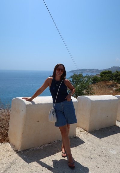 Shorts de mezclilla, blusa de tirantes azul marino y chanclas. Bolso ligero. De pie junto a una barrera de piedra blanca con un mar azul y un paisaje costero de fondo.