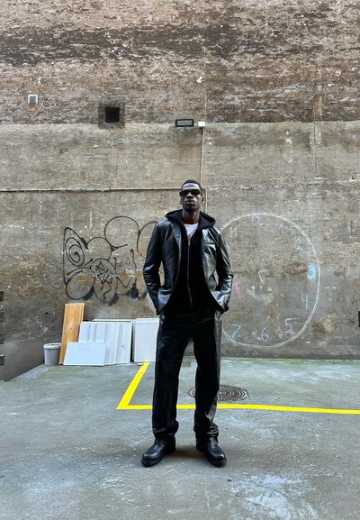 Chaqueta de cuero negra con capucha, pantalones a juego y botas negras gruesas. Gafas y un fondo texturizado con graffiti y un dibujo de un reloj.