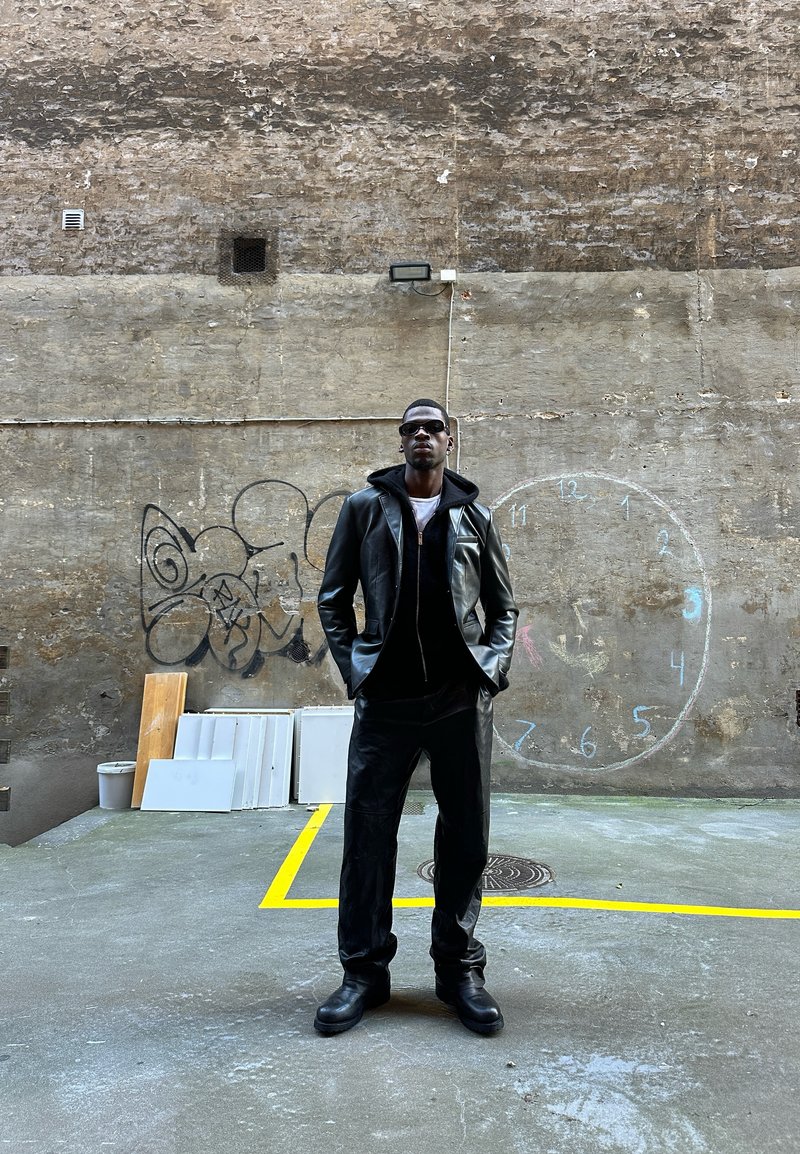 Schwarze Lederjacke mit Kapuze, passende Hose und klobige schwarze Stiefel. Brille und ein strukturierten Hintergrund mit Graffiti und einer Zeichnung einer Uhr.