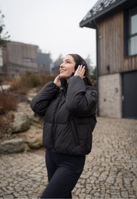 A parka preta possui um colarinho alto e fecho de zíper. Combinada com leggings pretas e protetores de orelha. O fundo inclui um caminho de pedras e um edifício de madeira.