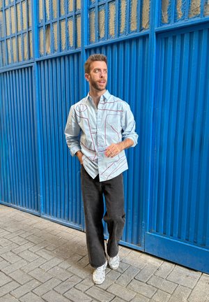 Camisa de botones de color azul claro con patrones geométricos de líneas rojas, combinada con pantalones negros de pierna ancha y zapatillas deportivas grises, sobre un pavimento de ladrillos.