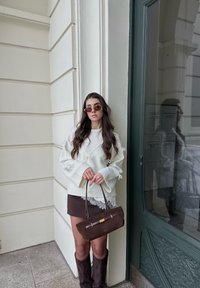 Femme aux longs cheveux portant des lunettes de soleil teintées, un pull blanc, une mini jupe marron avec de la dentelle, tenant un sac à main marron, debout près d'une porte verte et d'un mur blanc.