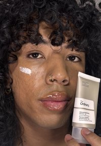 Putkilo "The Ordinary" kosteuttajasta pidetään kädessä, harmaassa, matta-pintaisessa pakkauksessa. Mallilla on kiharat hiukset, kostea iho ja valkoinen tuoteepäilys poskella.