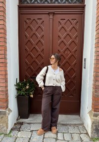 Cardigan en maille crème avec de gros boutons, pantalon rayé marron et chaussures couleur tan. Debout à côté d'une plante en pot près d'une porte marron.