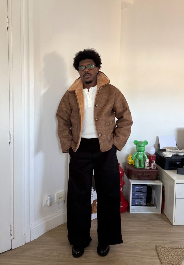 Man met een bril, gekleed in een bruine schapenvachtjas, wit overhemd en zwarte wijd uitlopende broek, staat binnen bij een tafel met decoratieve voorwerpen.