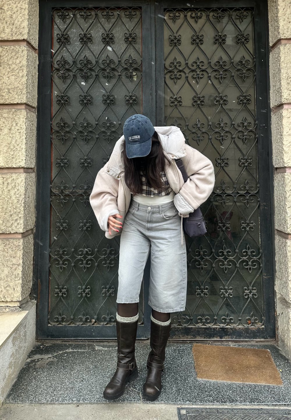 Cream oversized jacket, checked top, grey wide-leg shorts, and brown knee-high boots. Black cap and bag. Decorative black iron gate in the background.