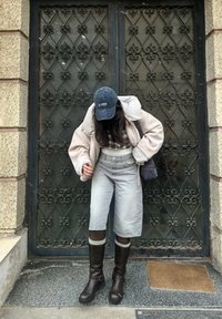 Cream oversized jacket, checked top, grey wide-leg shorts, and brown knee-high boots. Black cap and bag. Decorative black iron gate in the background.