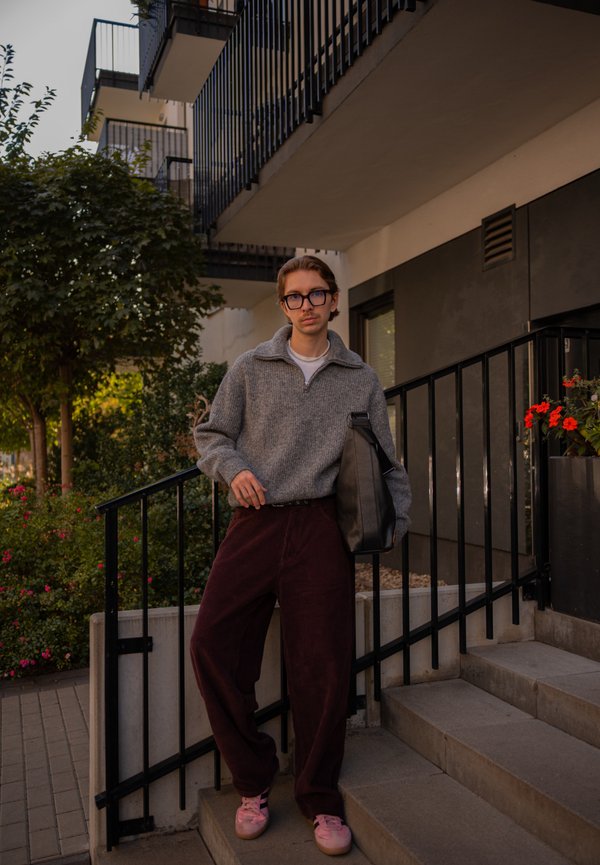 Un hombre con gafas, un jersey gris, pantalones burdeos y zapatos rosas está de pie en unos escalones exteriores con un bolso negro al hombro.