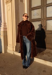 Femme portant un manteau en fourrure marron oversize, lunettes de soleil foncées, haut noir, jean bleu et chaussures noires à bout pointu, debout près de l'entrée d'un bâtiment.