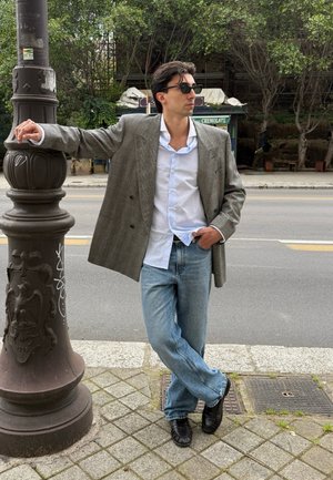 Hombre con blazer gris extragrande, camisa blanca, vaqueros azules y zapatos negros apoyado en un farol de la calle en una acera de la ciudad con árboles detrás.