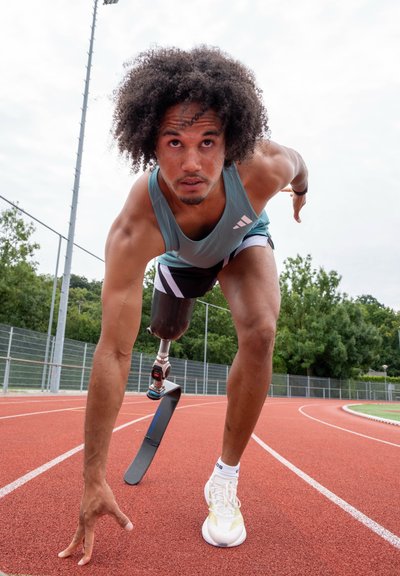 Atleta con una pierna protésica en posición de salida en una pista de atletismo al aire libre, vistiendo ropa deportiva y concentrado hacia adelante.