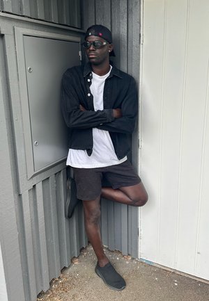 Chaqueta de mezclilla negra sobre una camiseta blanca, pantalones cortos oscuros y zapatos negros sin cordones. De pie contra una pared gris, con los brazos cruzados.