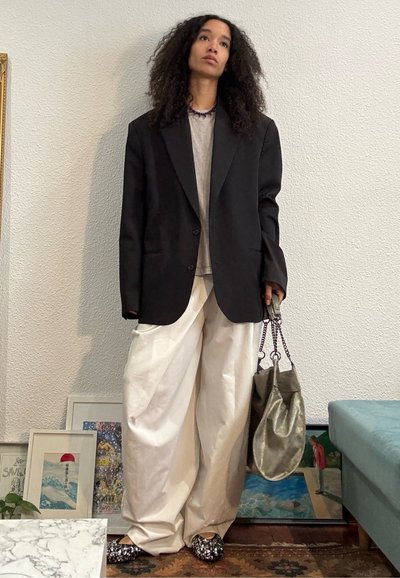 Persona con cabello rizado que lleva una chaqueta negra oversize, camisa beige, pantalones anchos blancos, sosteniendo un bolso metálico, de pie en interiores cerca de una obra de arte enmarcada.