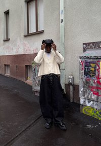 Fehér bolyhos kardigán fehér ing fölött, bő fekete farmernadrággal és vaskos fekete cipővel párosítva. Kiegészítők: szemüveg és fekete kalap.