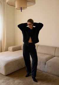 Melna, pūkaina, īsa kardigāna virs uzvalka, kombinēta ar augsta vidukļa tumšām džinsēm un melniem augstpapēžu apaviem ar smailiem purngalājiem. Fona mīkstajos toņos ir neitrāla dīvāna.