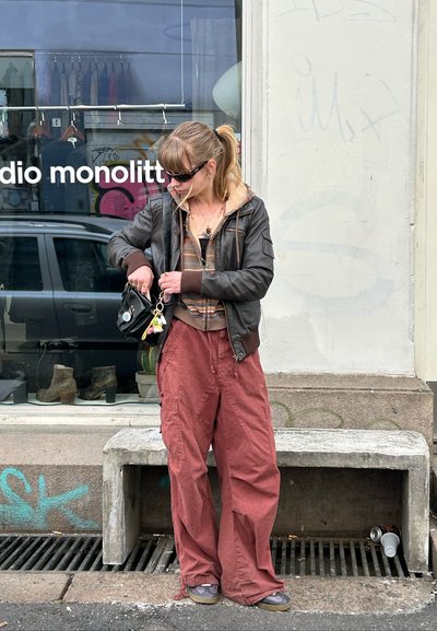 Persona con chaqueta de cuero marrón, camiseta de manga larga a rayas y pantalones rojos holgados, ajustando una pequeña bolsa negra con detalles de charms.