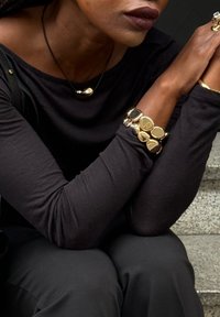 Gouden en zwarte sieraden omvatten een gladde gouden pendant ketting en een robuuste armband met diverse vormen van gouden en zwarte accenten tegen een zwarte top.