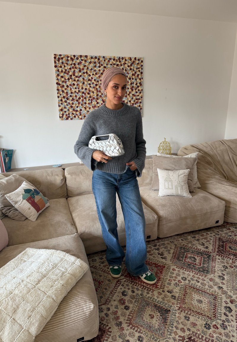 Frau mit beigem Kopftuch, grauem Pullover, blauen Jeans und grünen Turnschuhen steht in einem Wohnzimmer und hält eine weiße gewebte Handtasche.
