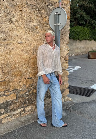 Camisa de lino de manga larga en color beige claro, con un corte holgado, combinada con unos pantalones vaqueros de pierna ancha en azul claro y chanclas, frente a una pared de piedra rústica.