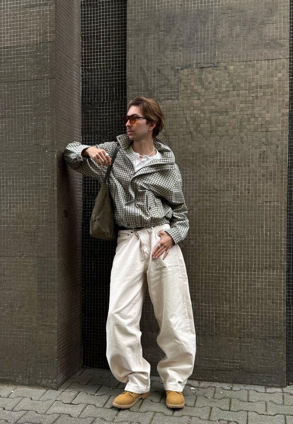 Una persona con gafas de sol, una chaqueta de cuadros, pantalones blancos holgados y zapatos de color tostado se apoya en una pared alicatada y lleva un bolso al hombro.
