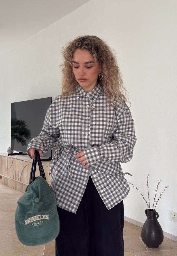 Jonge vrouw met krullend haar, gekleed in een grijs geruit overhemd en zwarte broek, met een groene tas in een minimalistische woonkamer.