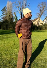Braune Pufferjacke mit einem Reißverschluss vorne und einer strukturierten Oberfläche, kombiniert mit beige hochgeschnittenen Hosen und einer cremefarbenen Mütze. Außensetting.