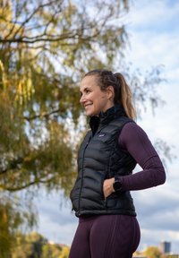 Vest puffer negru cu guler înalt, design ajustat, purtat peste o bluză cu mâneci lungi de culoare mov și colanți cu un model texturat.