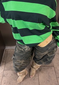 Zwart-groene gestreepte blouse met lange mouwen, camouflage cargoshorts en tan kleurige slouchy laarzen met open neuzen, die verschillende texturen en kleuren tonen.