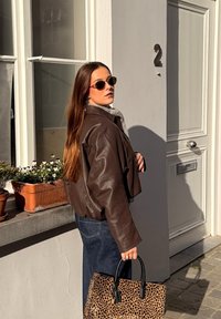 Femme aux longs cheveux portant une veste en cuir marron et des lunettes de soleil, tenant un sac à main à imprimé léopard, debout près d'une porte blanche numérotée 2.